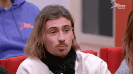 a man with long hair and a beard sits in a red chair in front of a screen that says star academy to live