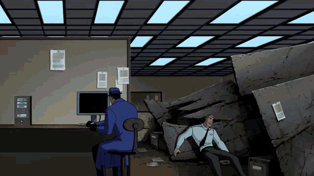 a man sits at a desk in front of a computer while another man lays on the floor