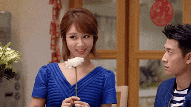 a woman in a blue dress is holding a white flower while a man looks on .