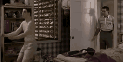 a man without a shirt is dancing in a bedroom with a man standing in the doorway .