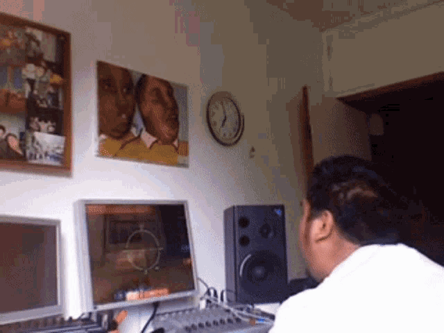 a man sits at a desk in front of two monitors and a clock with a picture of a couple on the wall behind him
