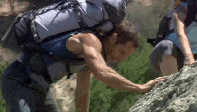 a man with a backpack is climbing a rock while a woman looks on .