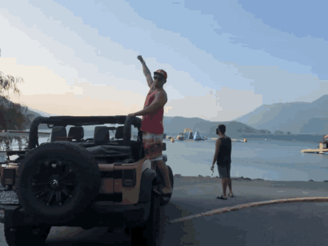 a man in a red shirt stands in front of a jeep with his fist in the air