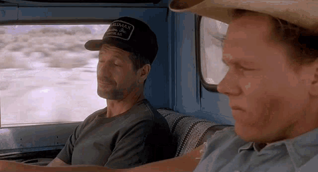 two men are sitting in the back seat of a truck and one of them is wearing a black hat that says klamath