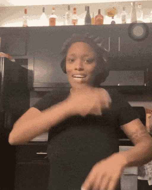 a woman in a black shirt is dancing in a kitchen with bottles on the shelf behind her