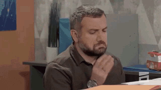 a man with a beard is sitting at a desk in an office with his hand on his chin .