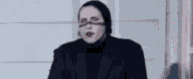 a woman wearing glasses and a black jacket is sitting in front of a white wall .
