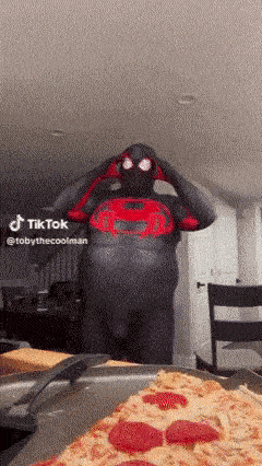 a man in a spider-man costume is standing next to a pizza .