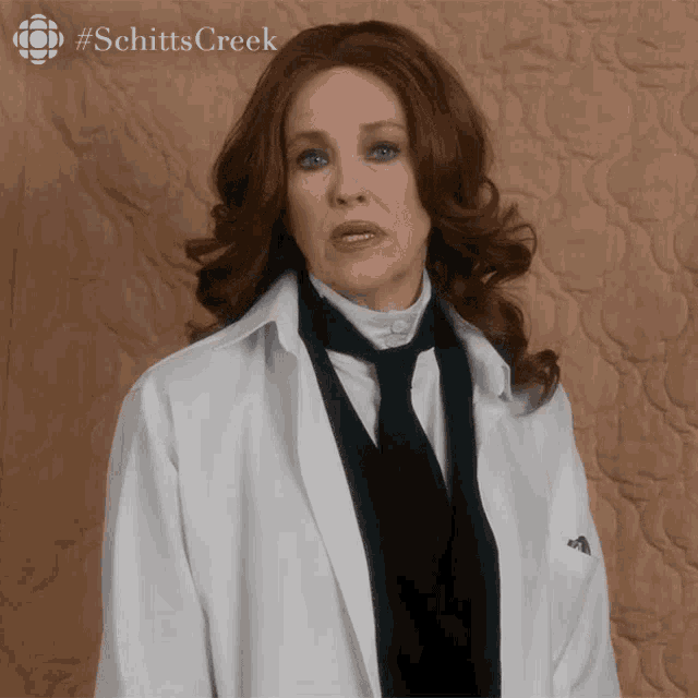 a woman in a white coat and black tie is standing in front of a quilt with #schitts creek written on the bottom