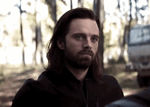 a man with long hair and a beard is standing in the woods looking at something .