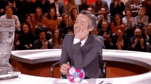 a man in a suit and tie is laughing in front of a crowd with tmc written on the bottom