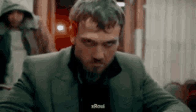 a man with a beard is sitting at a table and looking at the camera with a hooded man in the background .