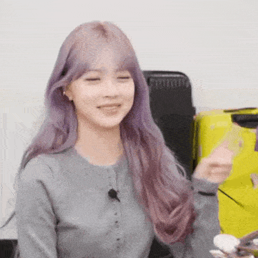 a woman with purple hair is sitting in front of a yellow suitcase and smiling .