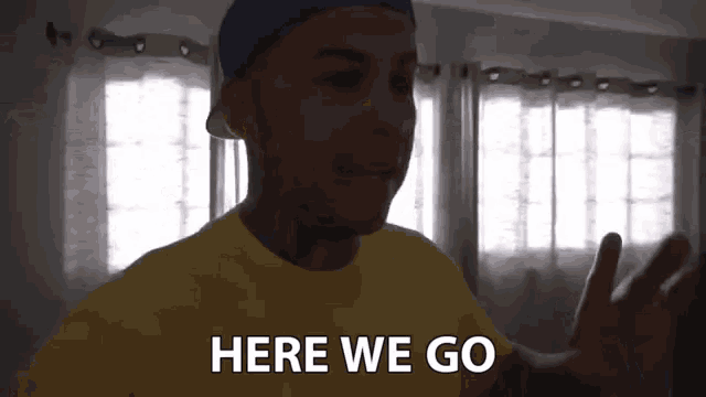 a man in a yellow shirt is standing in front of a window and says here we go