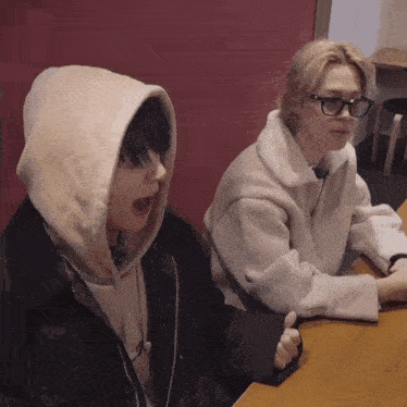 two people are sitting at a table with one wearing glasses and the other wearing a hoodie