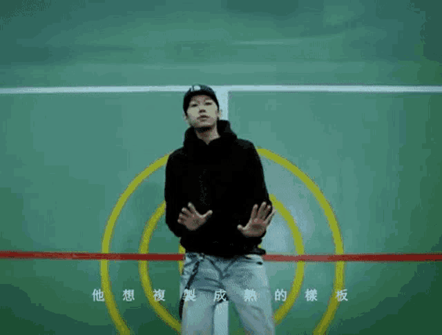 a man in a black hoodie is standing in front of a green wall with chinese writing on it .