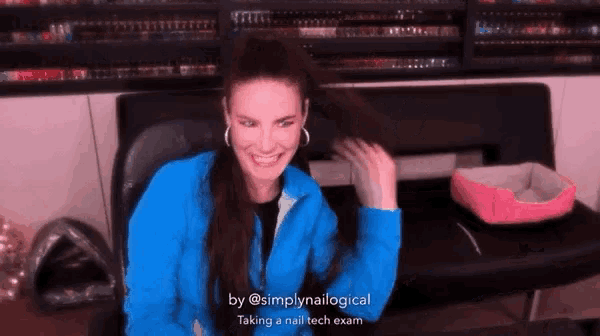 a woman in a blue jacket is sitting at a desk and smiling while taking a nail tech exam