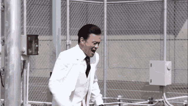 a man in a white suit and black tie stands in front of a chain link fence