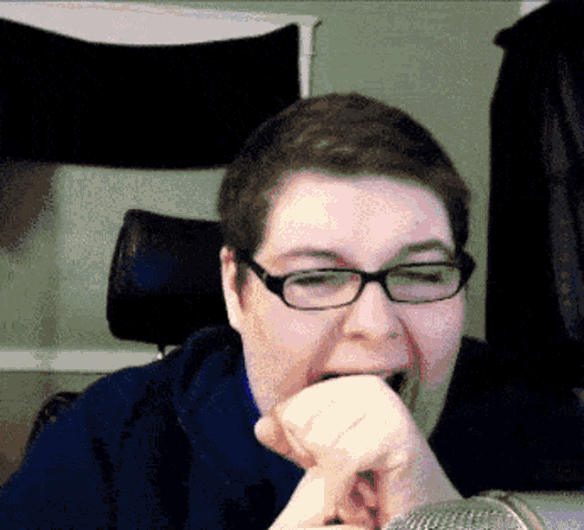 a man wearing glasses and a blue sweater is biting into a microphone