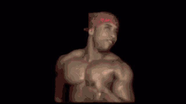 a shirtless man wearing a red bandana and a red hat is standing in the dark .