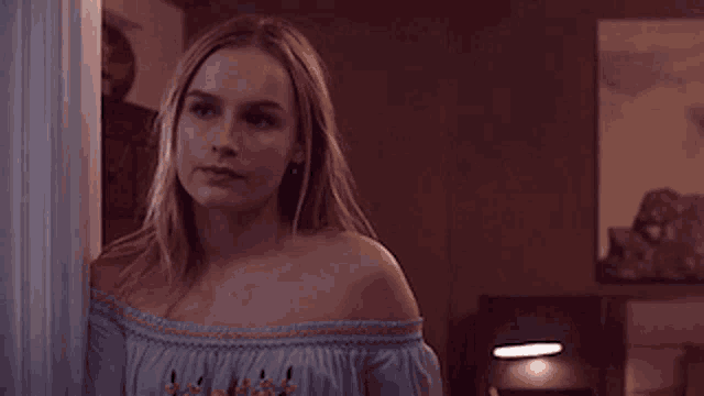 a woman in a blue off the shoulder top stands in a dark room .