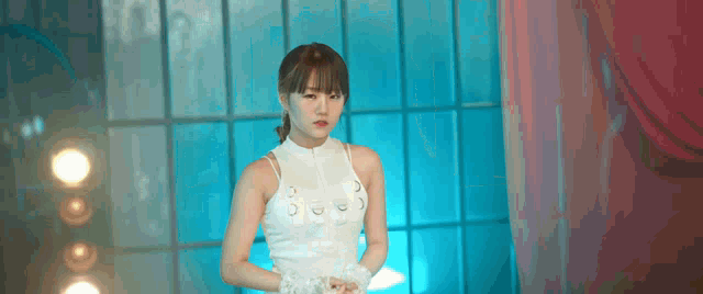 a woman in a white dress is standing in front of a window and looking at the camera .