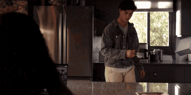 a man in a denim jacket stands in a kitchen holding a cup