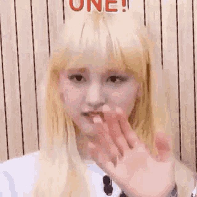 a close up of a woman with blonde hair waving her hand in front of a wooden wall .