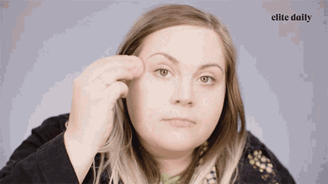 a woman applying makeup with a sponge with the elite daily logo behind her