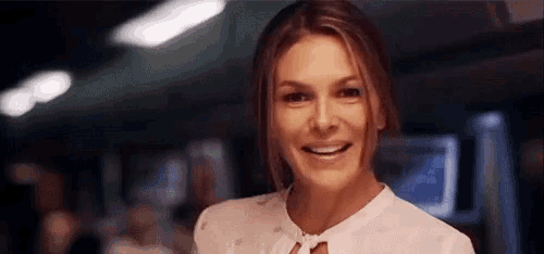 a woman in a white shirt is smiling and looking at the camera in a dark room .