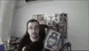 a blurry picture of a man holding a book in front of a wall with pictures on it .
