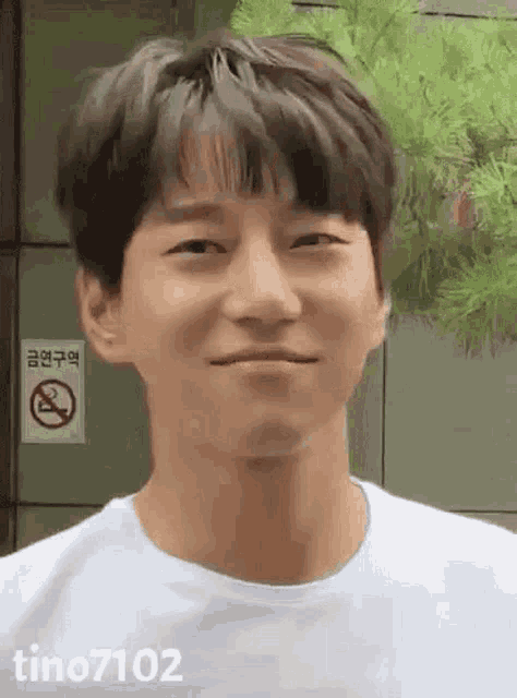 a young man wearing a white t-shirt is smiling in front of a no smoking sign .