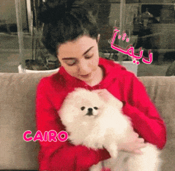 a woman in a red hoodie is holding a small white dog with the name cairo on the bottom
