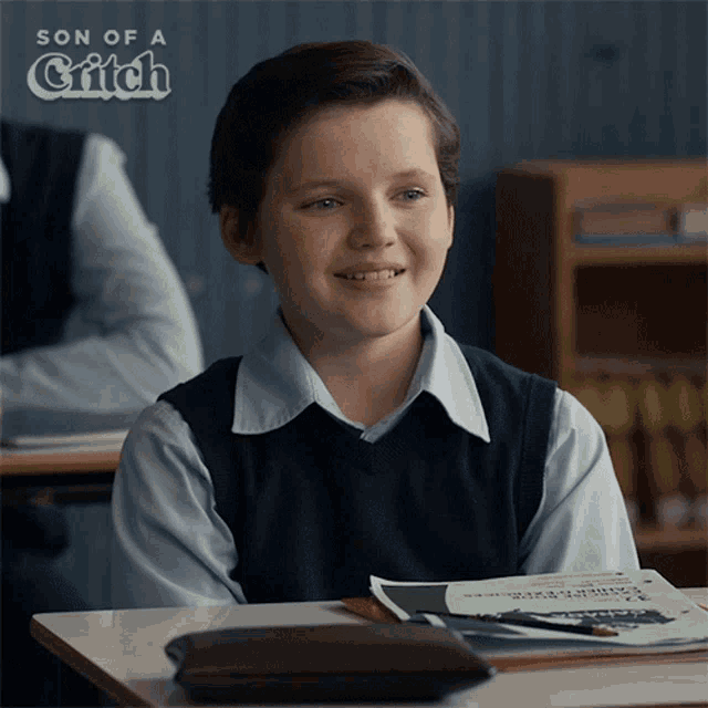 a young boy sits at a desk in a classroom with a son of a grifch advertisement behind him