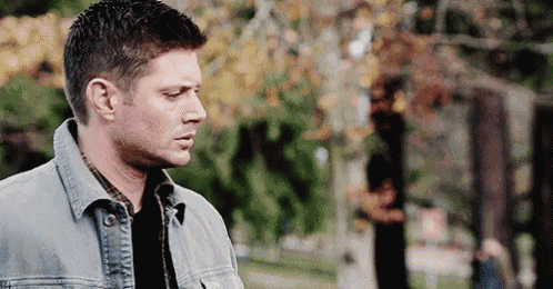 a man in a denim jacket is standing in a park looking down .