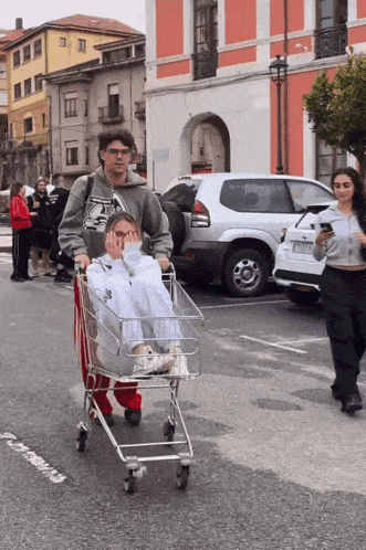 a man wearing a sweatshirt that says ' reebok ' on it pushes a woman in a shopping cart down a street