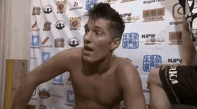 a man without a shirt is standing in front of a wall covered in logos .