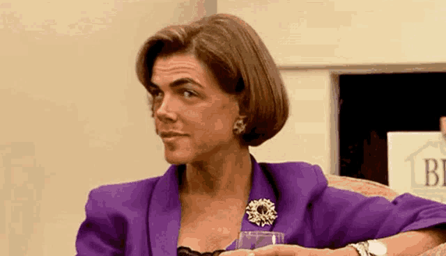 a woman in a purple suit and brooch is sitting on a couch holding a glass of wine .