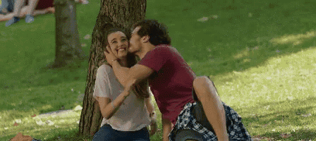 a man is kissing a woman on the cheek while sitting under a tree in the park .