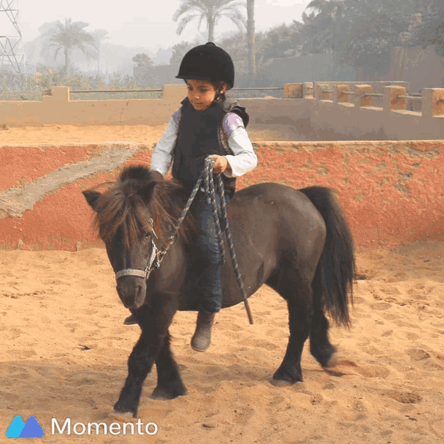 a little girl is riding a pony in the sand with the momento logo behind her