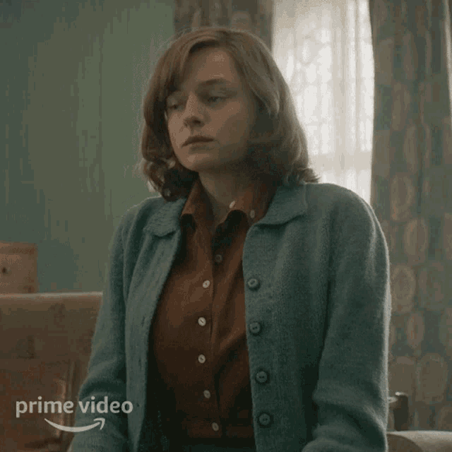 a woman wearing a blue cardigan is sitting in front of a window with a prime video logo on the bottom