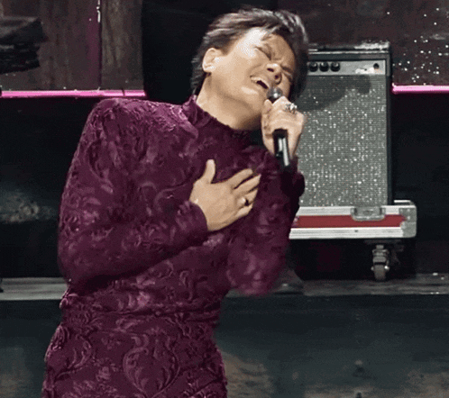 a woman in a purple dress is singing into a microphone with her hands on her chest