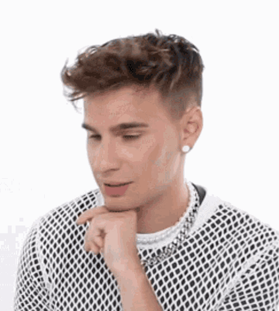 a young man wearing a fishnet shirt and a pearl necklace is looking down with his hand on his chin .
