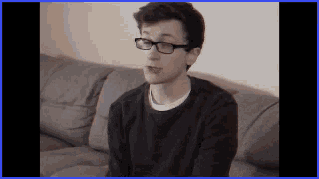 a young man wearing glasses and a black sweater is sitting on a couch