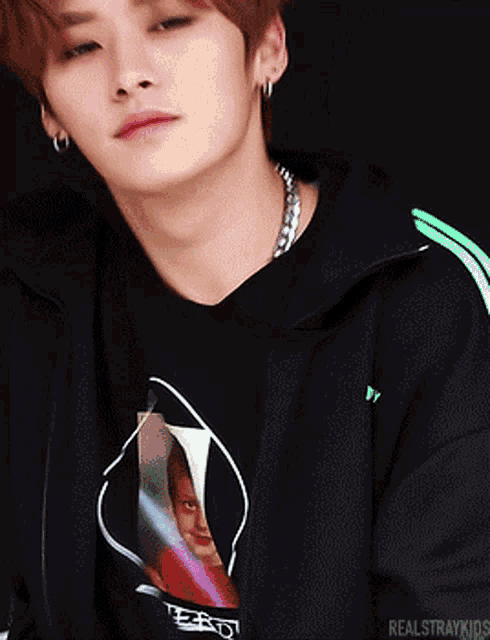 a young man wearing a black t-shirt and a black jacket with green stripes is looking at the camera .