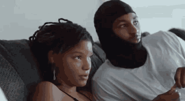 a man and a woman are sitting on a couch looking at something . the man is wearing a mask .