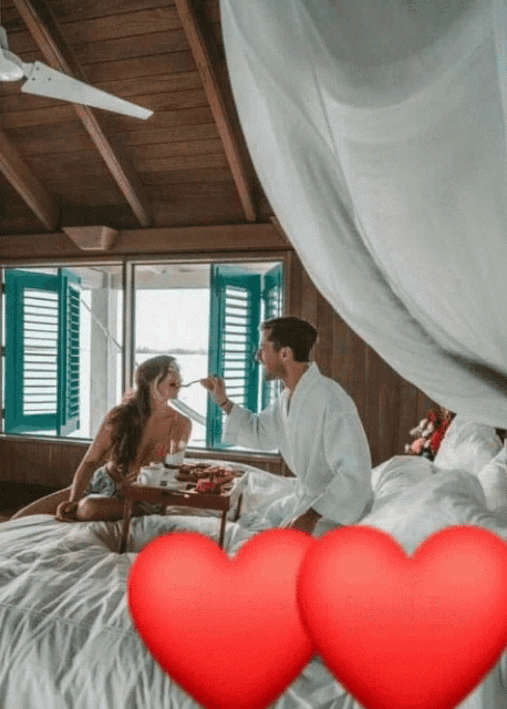 a man feeding a woman on a bed with two red hearts in the background