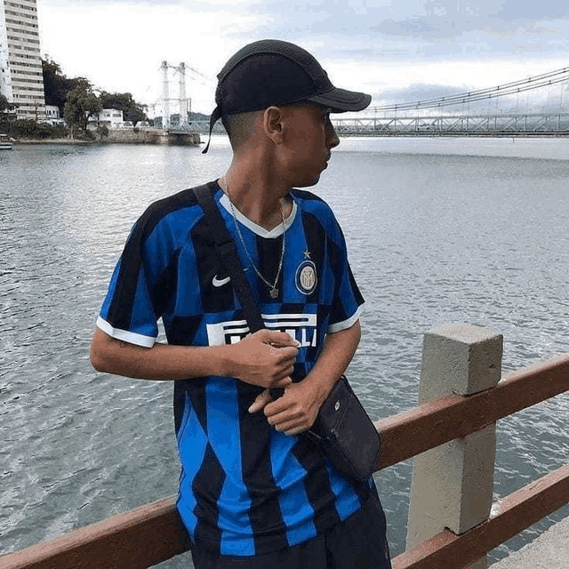 a young man in a blue and black striped shirt is standing on a bridge overlooking a river .