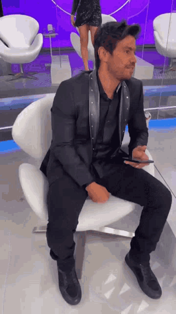a man in a black suit is sitting in a white chair holding a cell phone