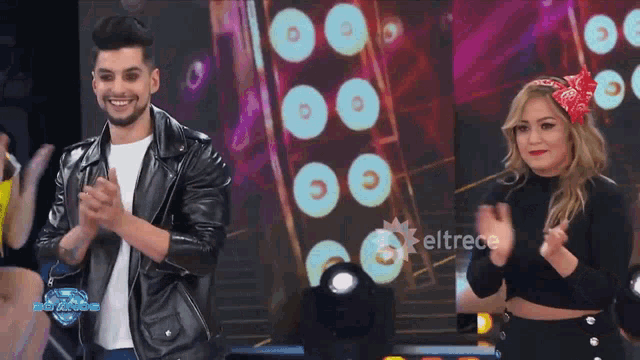 a man in a leather jacket and a woman in a crop top are clapping in front of a sign that says eltrece